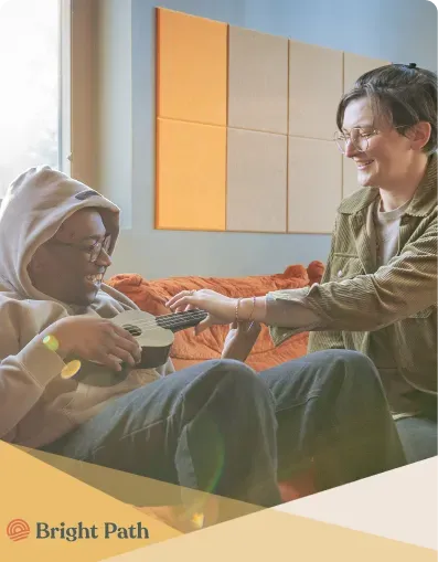 Young person playing a ukulele on an orange couch while a woman leans in at Bright Path