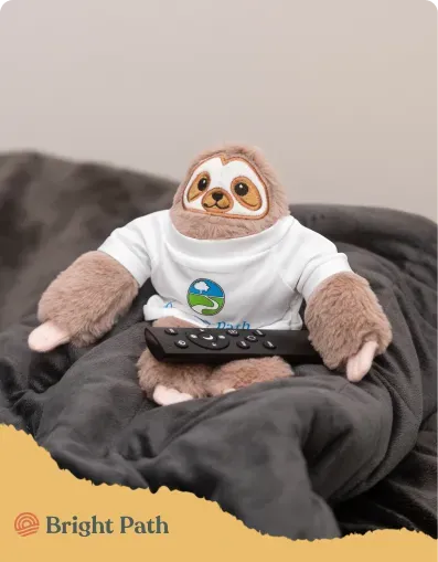 Bright Path sloth plush holding a TV remote on a gray blanket