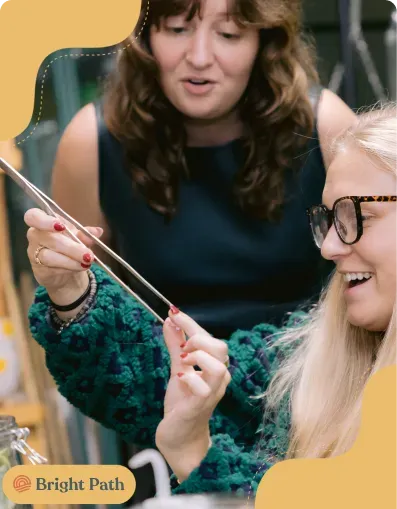 Woman using long tweezers on a terrarium as another woman laughs at Bright Path