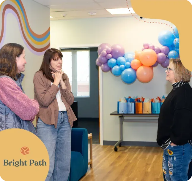 Three women talking beside a balloon arch and gift bags in a Bright Path lounge with a rainbow wave mural